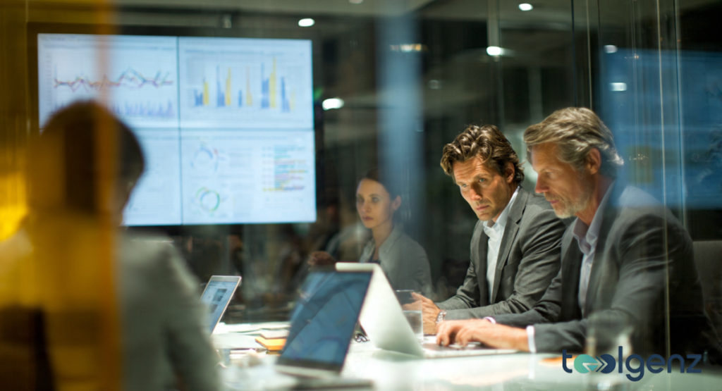Professional business meeting in a modern glass-walled conference room featuring executives looking at laptops and large data visualization screens.