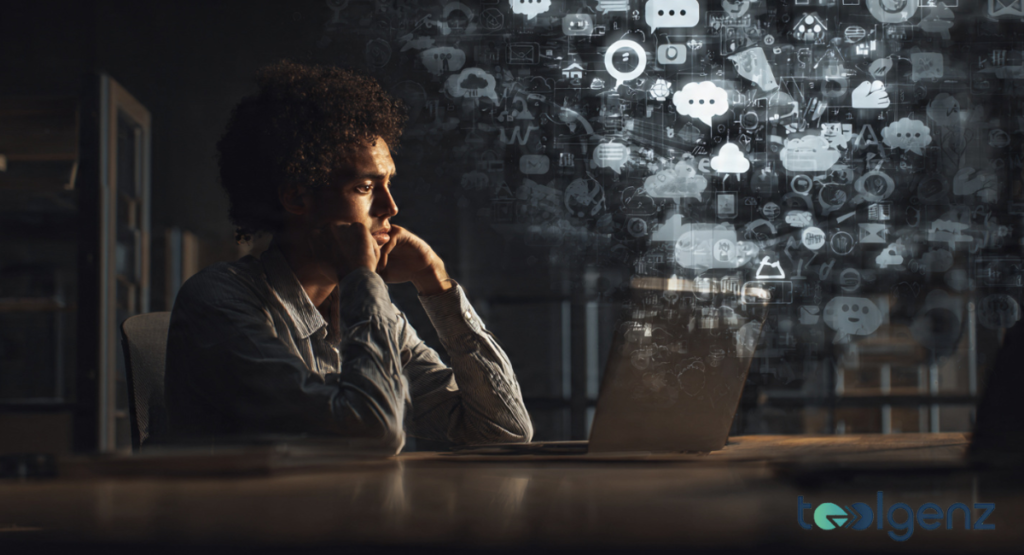 A man sitting at a laptop in a dark room, looking stressed by a cloud of digital icons. The image represents information overload and ethical fatigue.